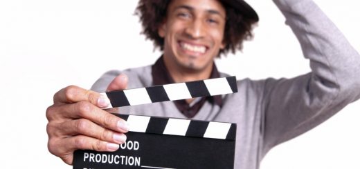 A smiling man wearing a hat holds a film clapperboard toward the camera.