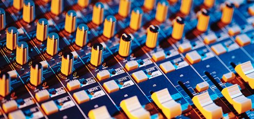Close-up of an audio mixing console with knobs, buttons, and faders illuminated in orange and blue light.
