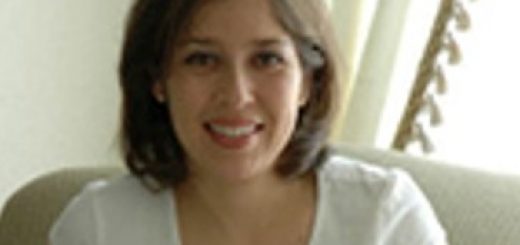 A smiling woman with shoulder-length brown hair in a white shirt sits on a couch.