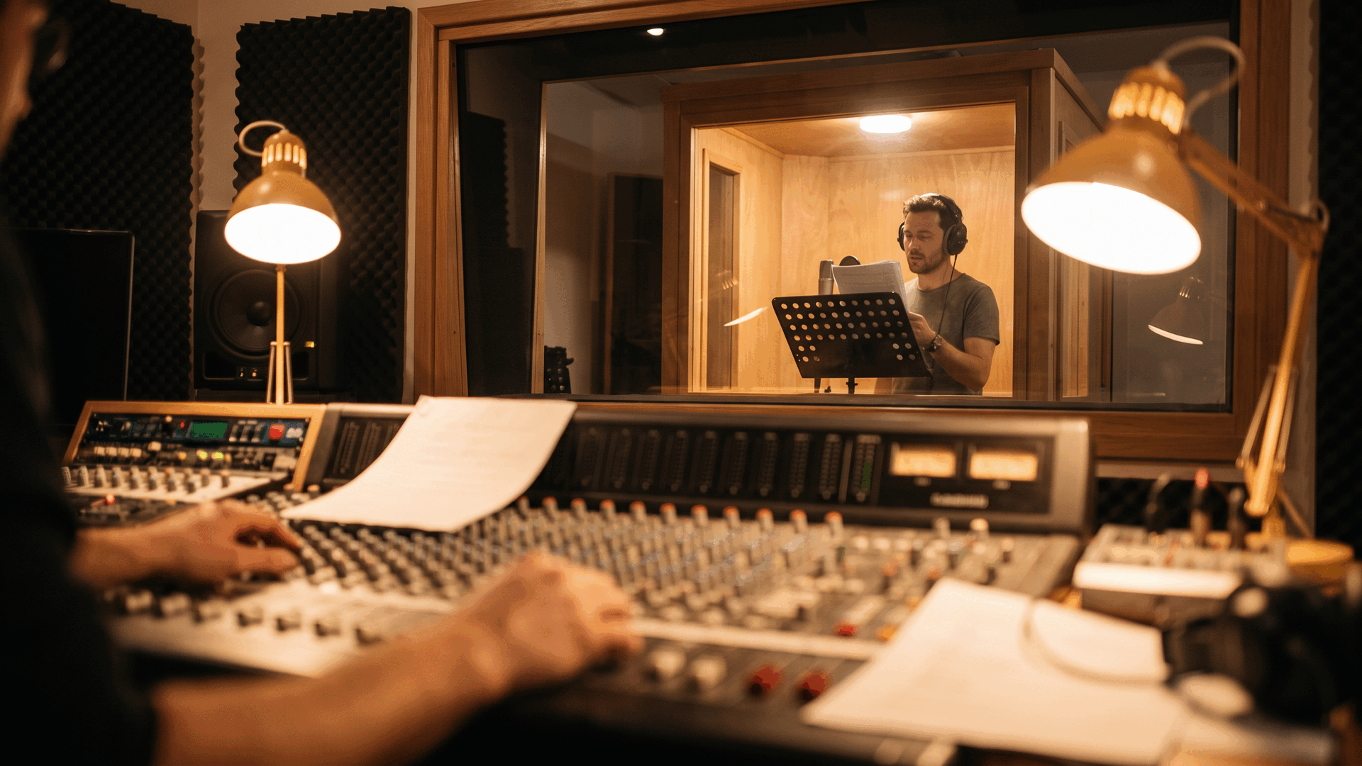 A sound engineer adjusts a mixing console while a man in headphones reads from a script in a vocal booth.