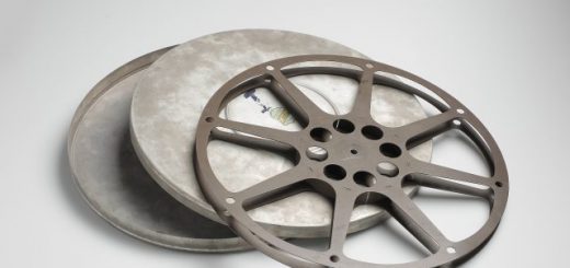 A metal film reel rests on an open round film canister against a plain background.