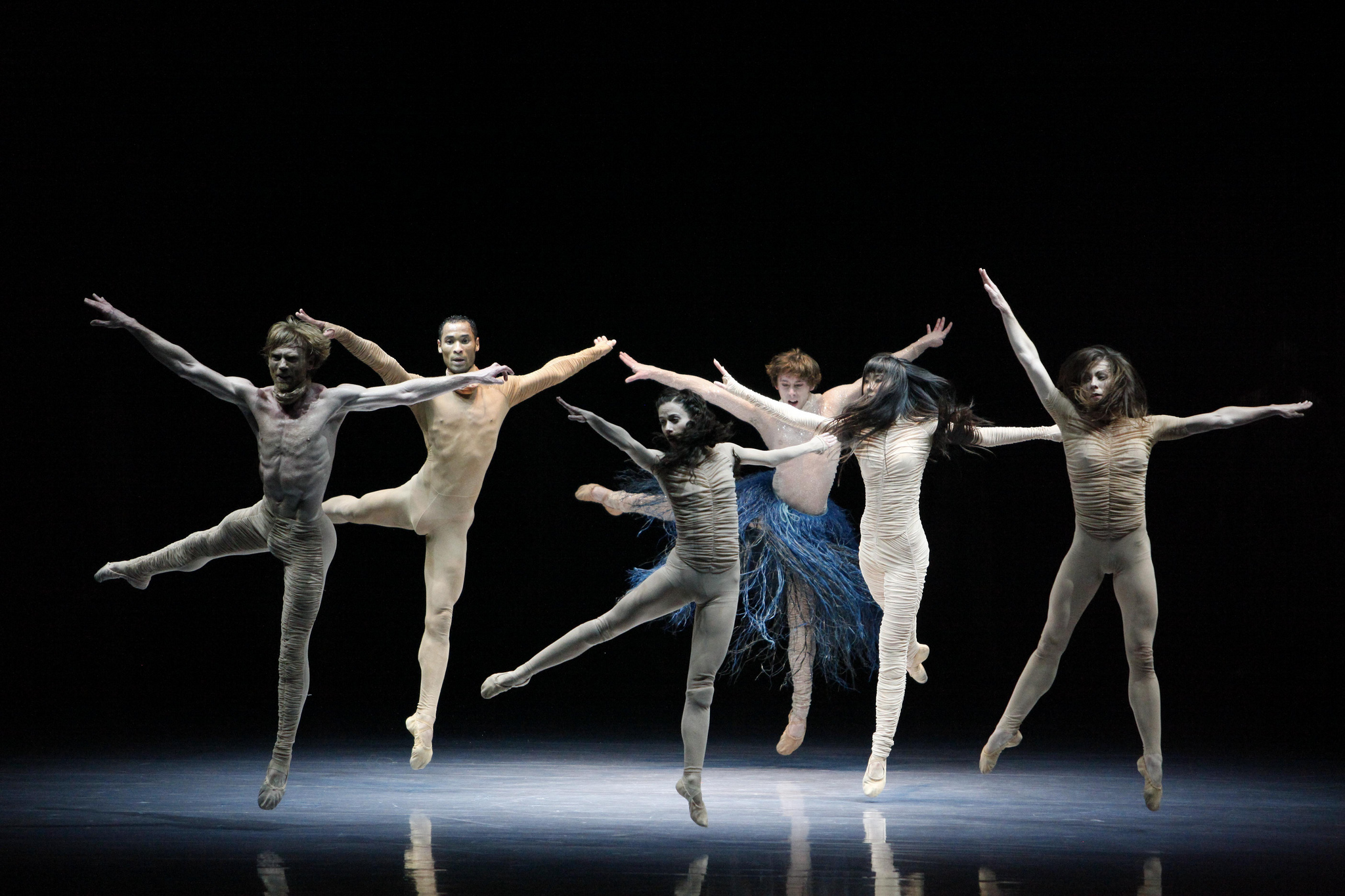 Six dancers in neutral bodysuits leap across a dark stage with arms outstretched.