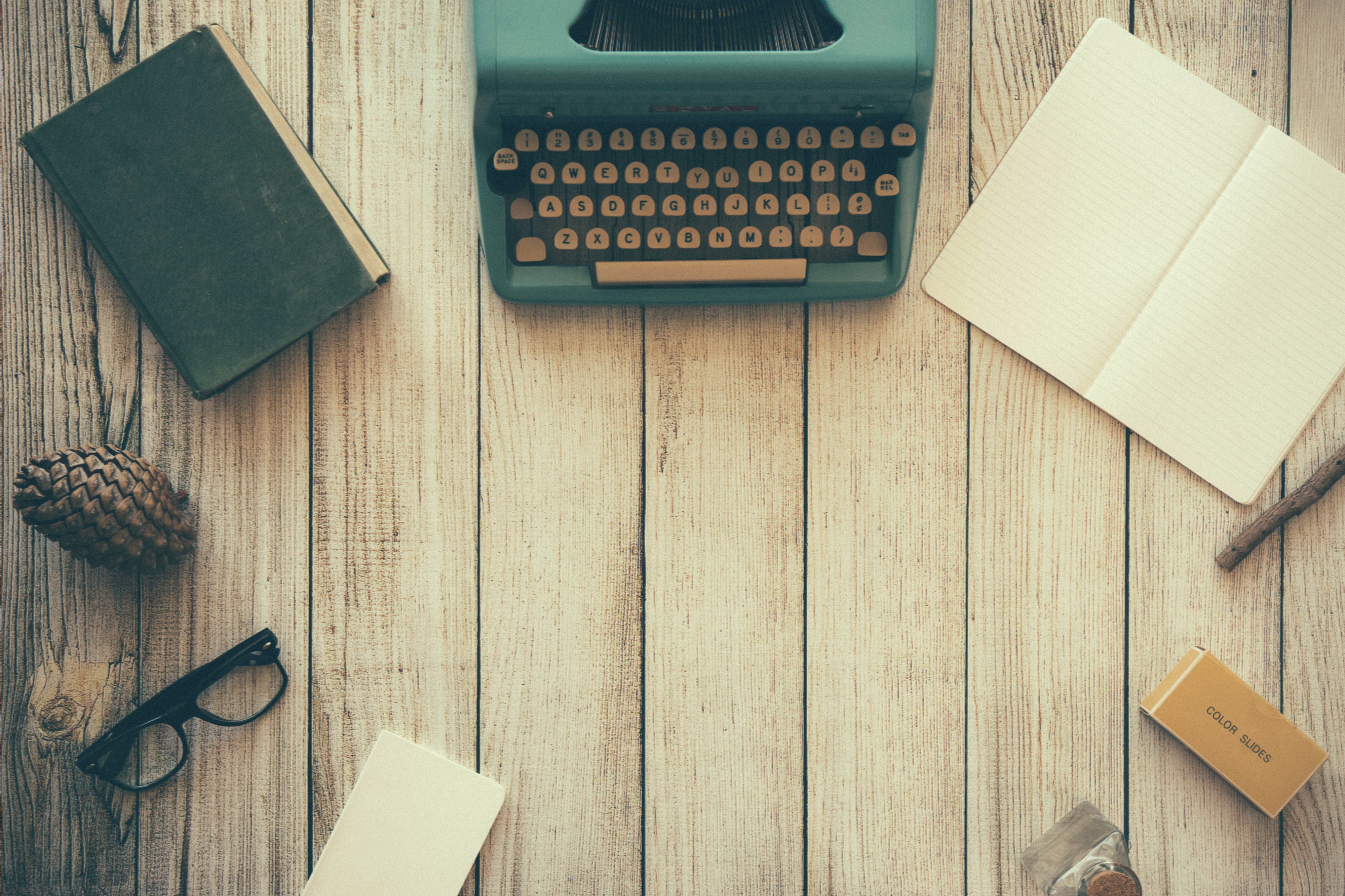 Vintage typewriter, open notebook, book, and eyeglasses arranged around empty space on a wooden desk.
