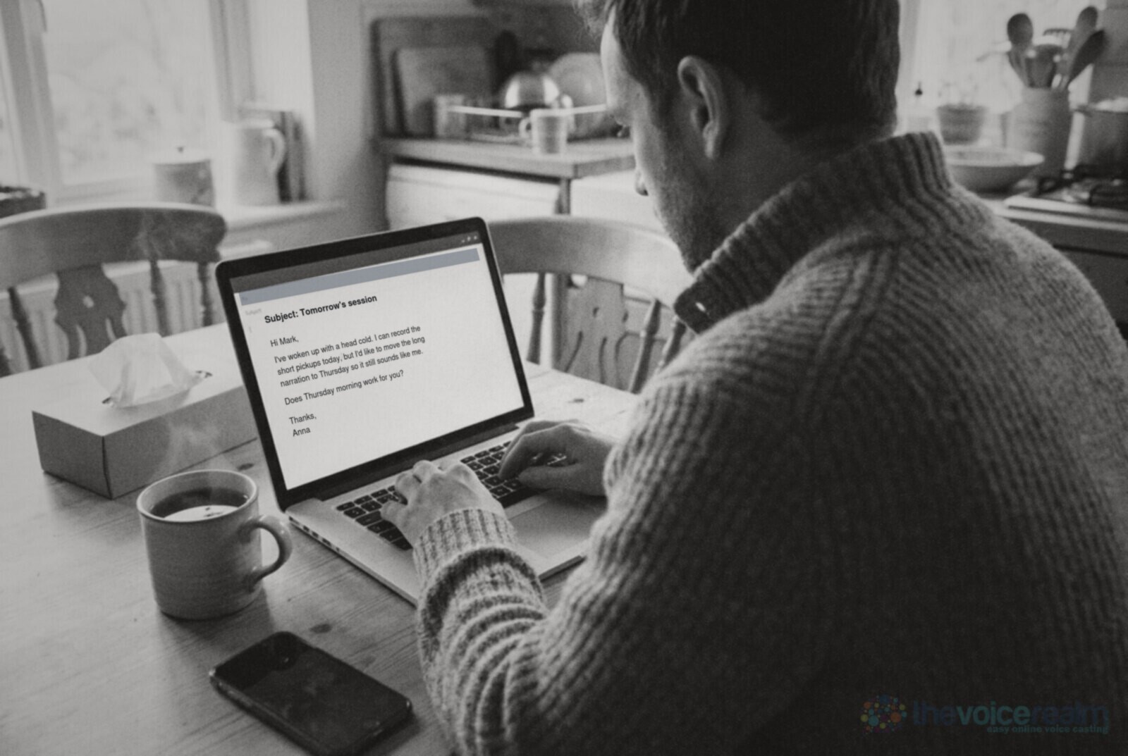 Over-the-shoulder view of a man in a wool jumper typing an email at a kitchen table, a mug of tea, a box of tissues and a phone lying face down beside the laptop © The Voice Realm
