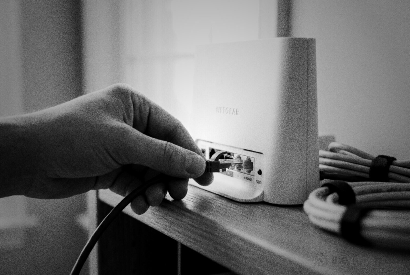 A hand pushing a blue ethernet cable into the back of a white home wifi router on a wooden shelf © The Voice Realm
