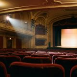 An empty theatre auditorium seen from the back of the stalls, rows of red velvet seats and a softly lit empty stage in the distance © The Voice Realm