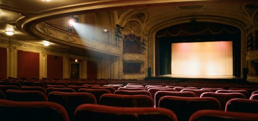 An empty theatre auditorium seen from the back of the stalls, rows of red velvet seats and a softly lit empty stage in the distance © The Voice Realm
