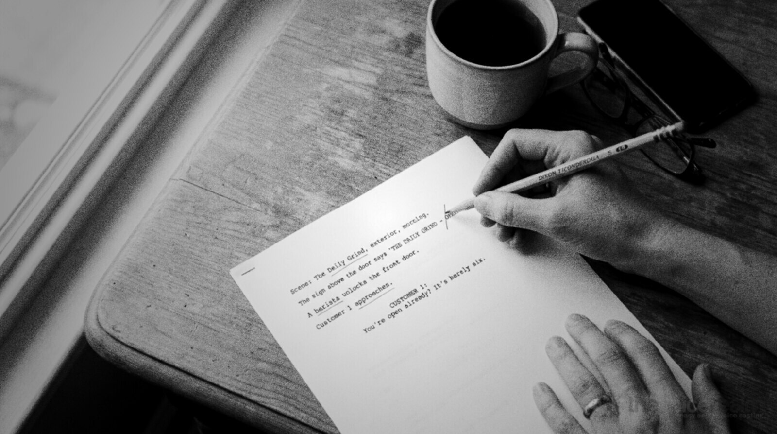 Overhead view of a printed script on a wooden kitchen table, a hand drawing a pencil slash between two words, a mug of tea, folded reading glasses and a phone beside it © The Voice Realm