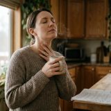 A voice actor at her kitchen window in the morning, eyes closed, humming gently with a hand on her throat and a mug of tea, a marked-up script on the counter