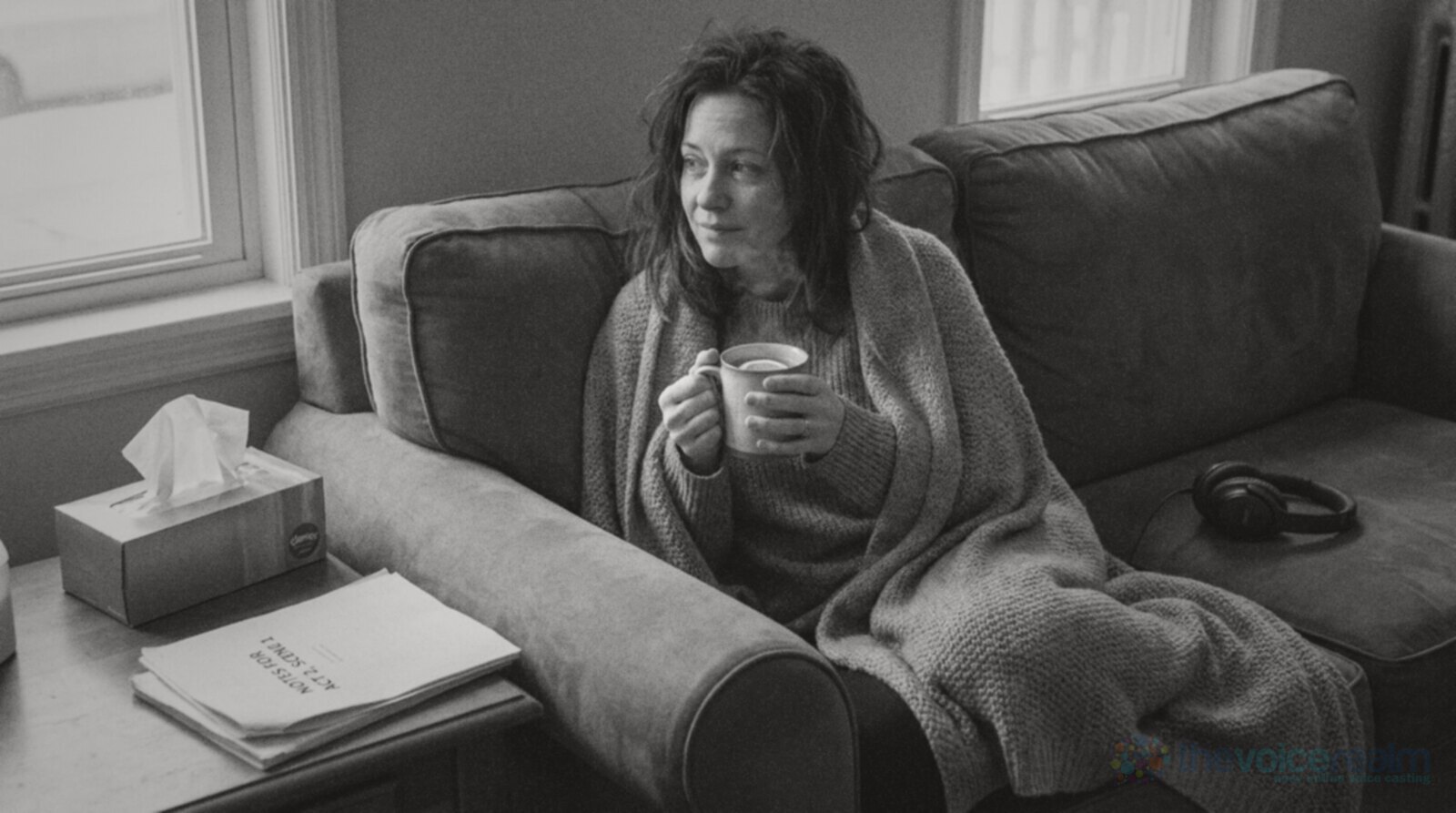 A tired woman wrapped in a grey blanket on a sofa, holding a mug of hot water with lemon, a box of tissues and a printed script on the side table and closed-back headphones resting on the sofa beside her © The Voice Realm