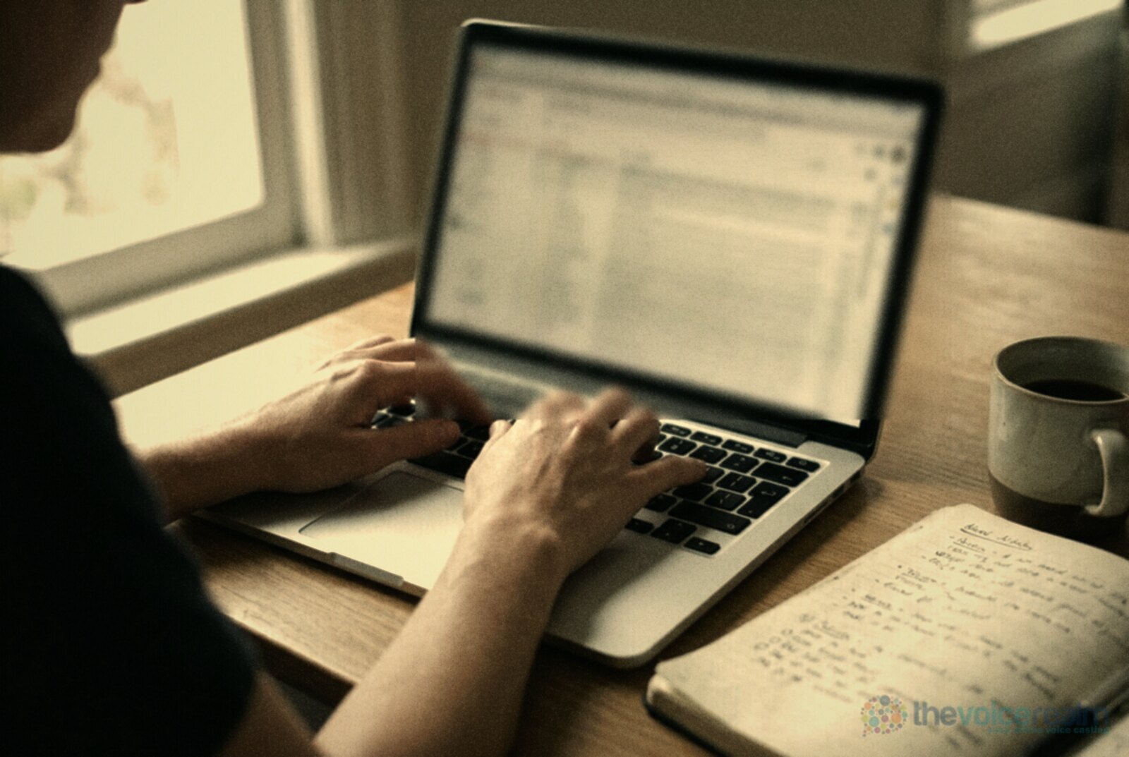 A person typing a reply on a laptop at a wooden table, a notebook of handwritten notes and a mug of coffee beside it © The Voice Realm