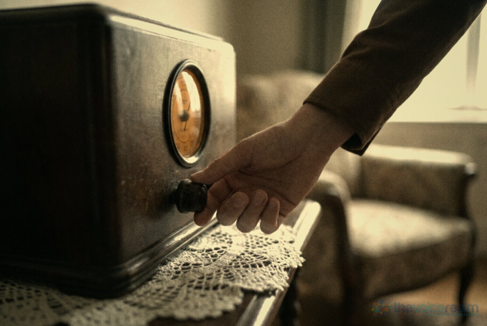 A hand turning the tuning knob of an antique wooden valve radio with a glowing dial, on a lace doily in a sitting room © The Voice Realm