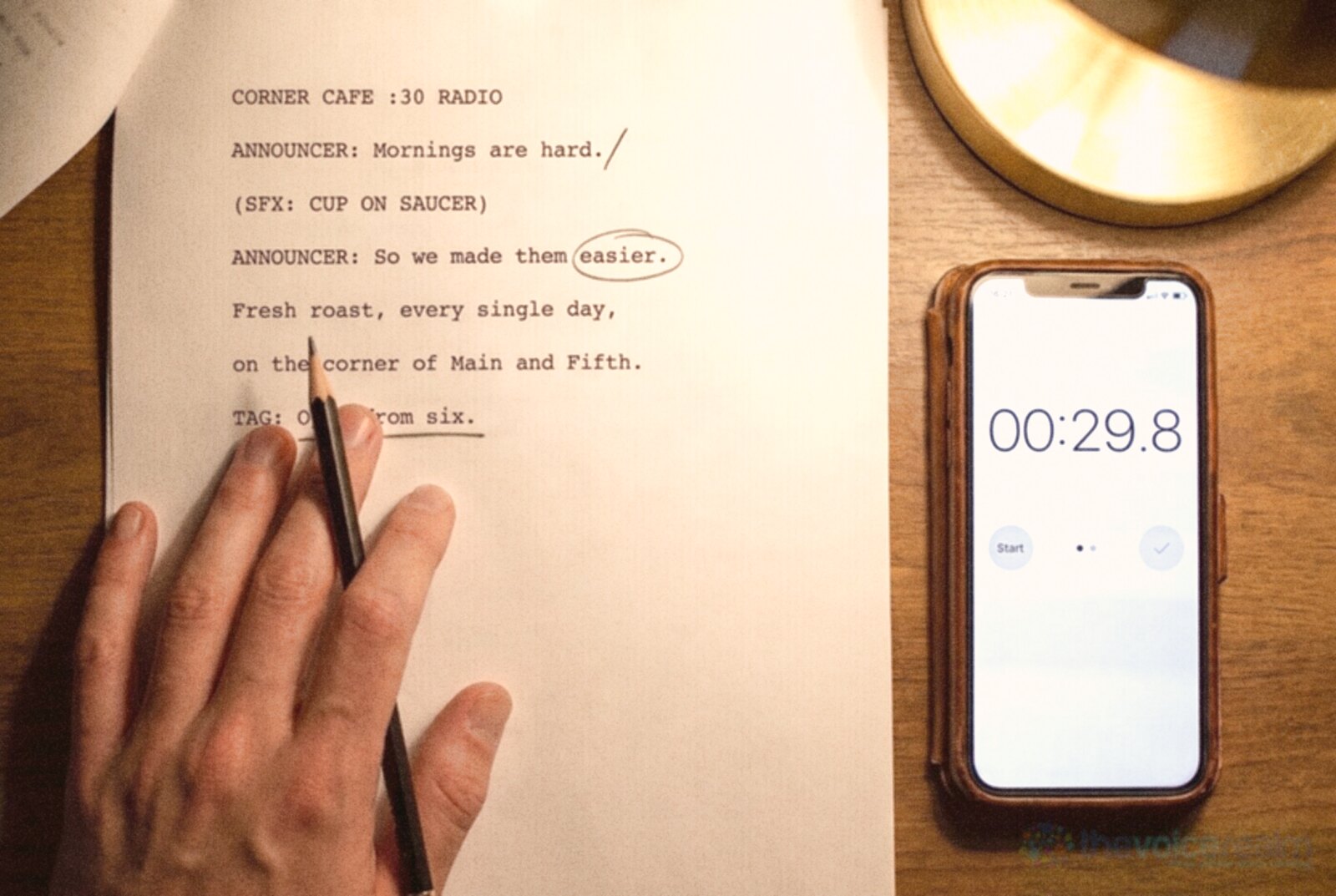 A printed script marked up in pencil with a circled word, slash marks and an arrow, a hand holding the pencil and a phone stopwatch beside it © The Voice Realm