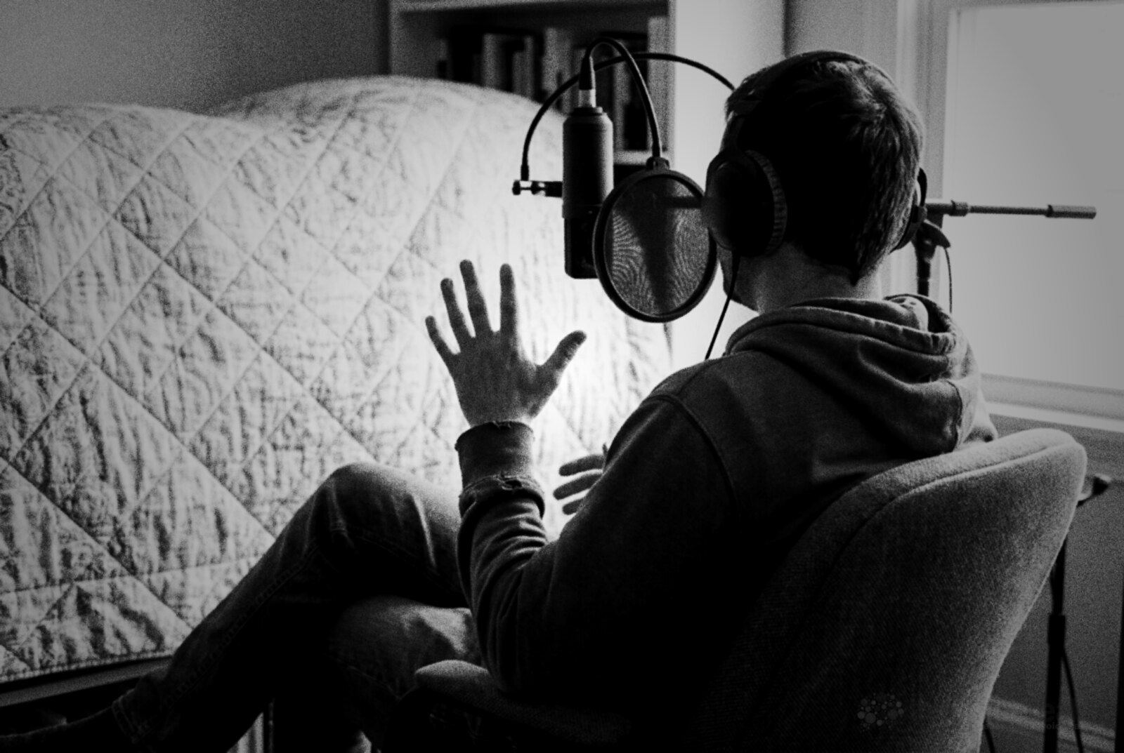 A voice actor seen from behind in headphones, relaxed in a chair at a microphone with a pop filter, one hand raised mid-gesture as if chatting © The Voice Realm