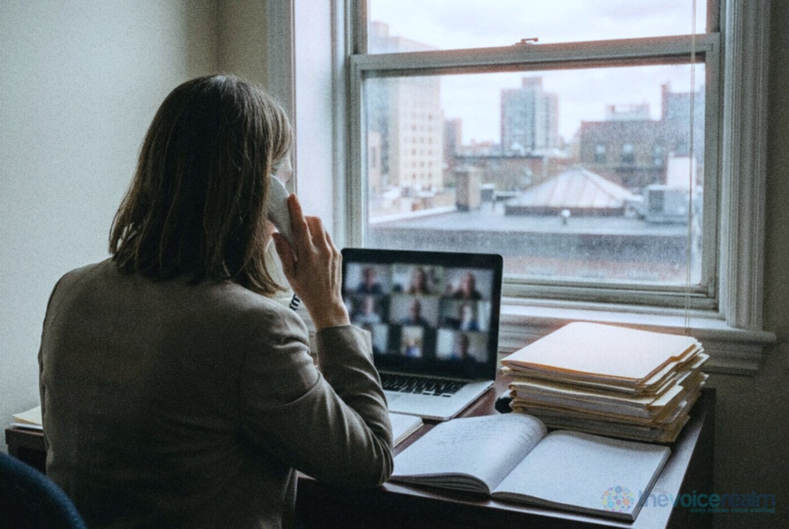 A woman seen from behind on a desk phone call at a laptop in a city office, a stack of folders beside her © The Voice Realm