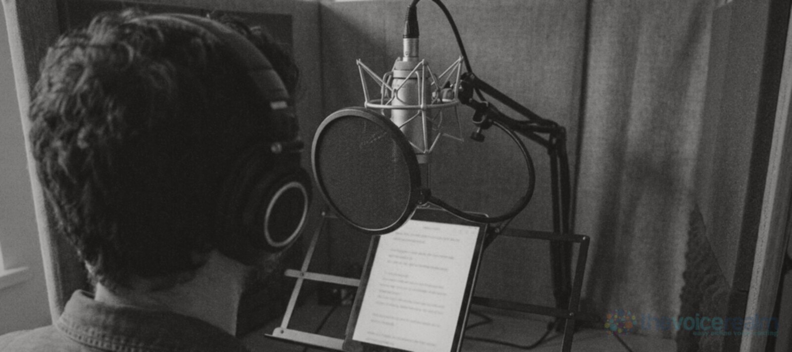 A voice actor in headphones seen from behind at a condenser microphone set a little above mouth height, a round pop filter in front of it and a tablet script on a stand below © The Voice Realm