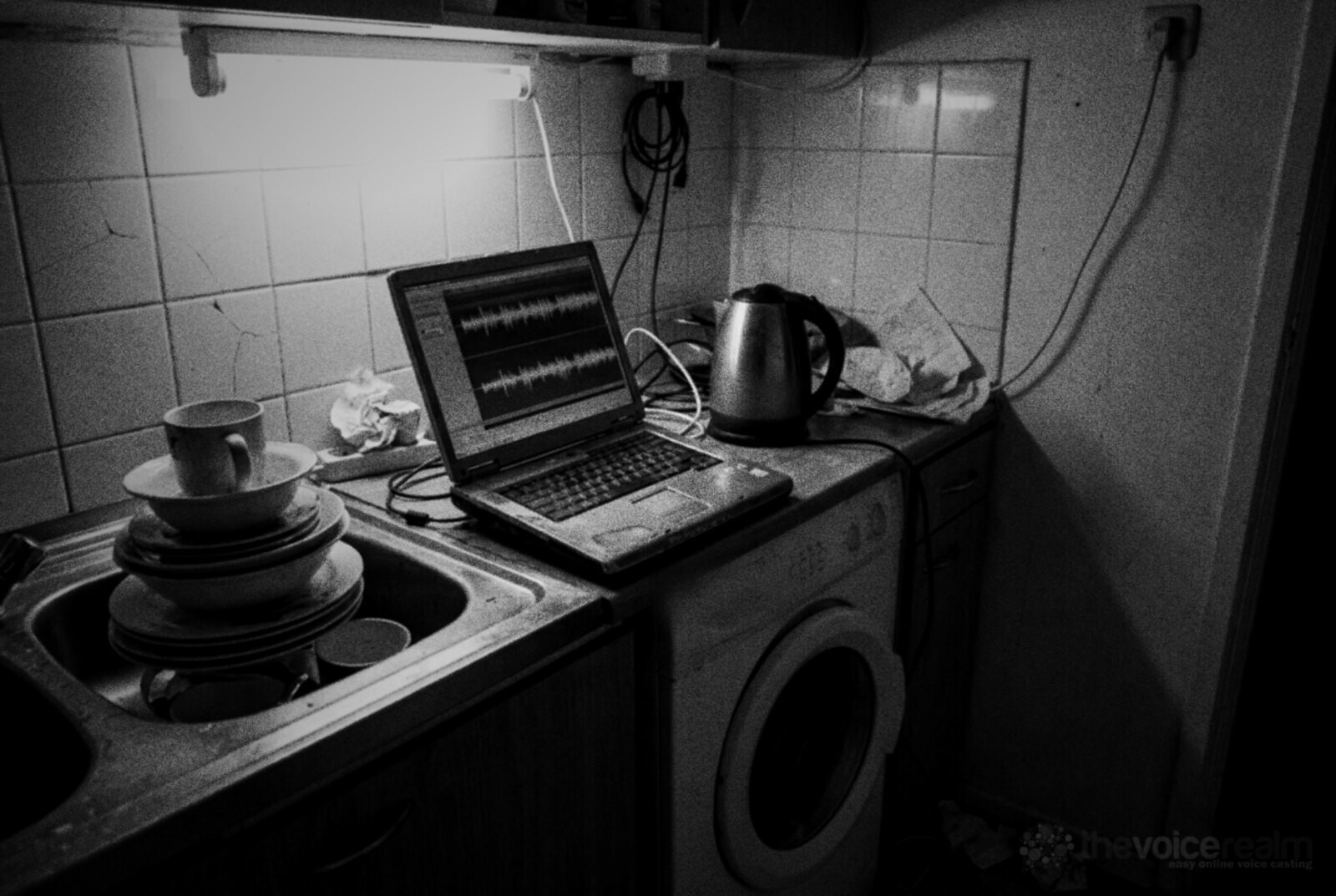 An old laptop showing an audio waveform on a cluttered kitchen bench beside a washing machine, a kettle and a sink of dishes © The Voice Realm