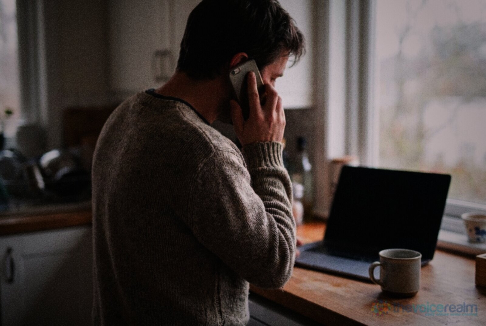 A man in a knitted jumper stands at a kitchen bench with a phone to his ear, a laptop and a mug beside him © The Voice Realm
