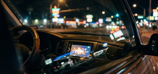 Inside a car at night, the dashboard display glowing, with the blurred neon lights of a city strip beyond the windscreen © The Voice Realm