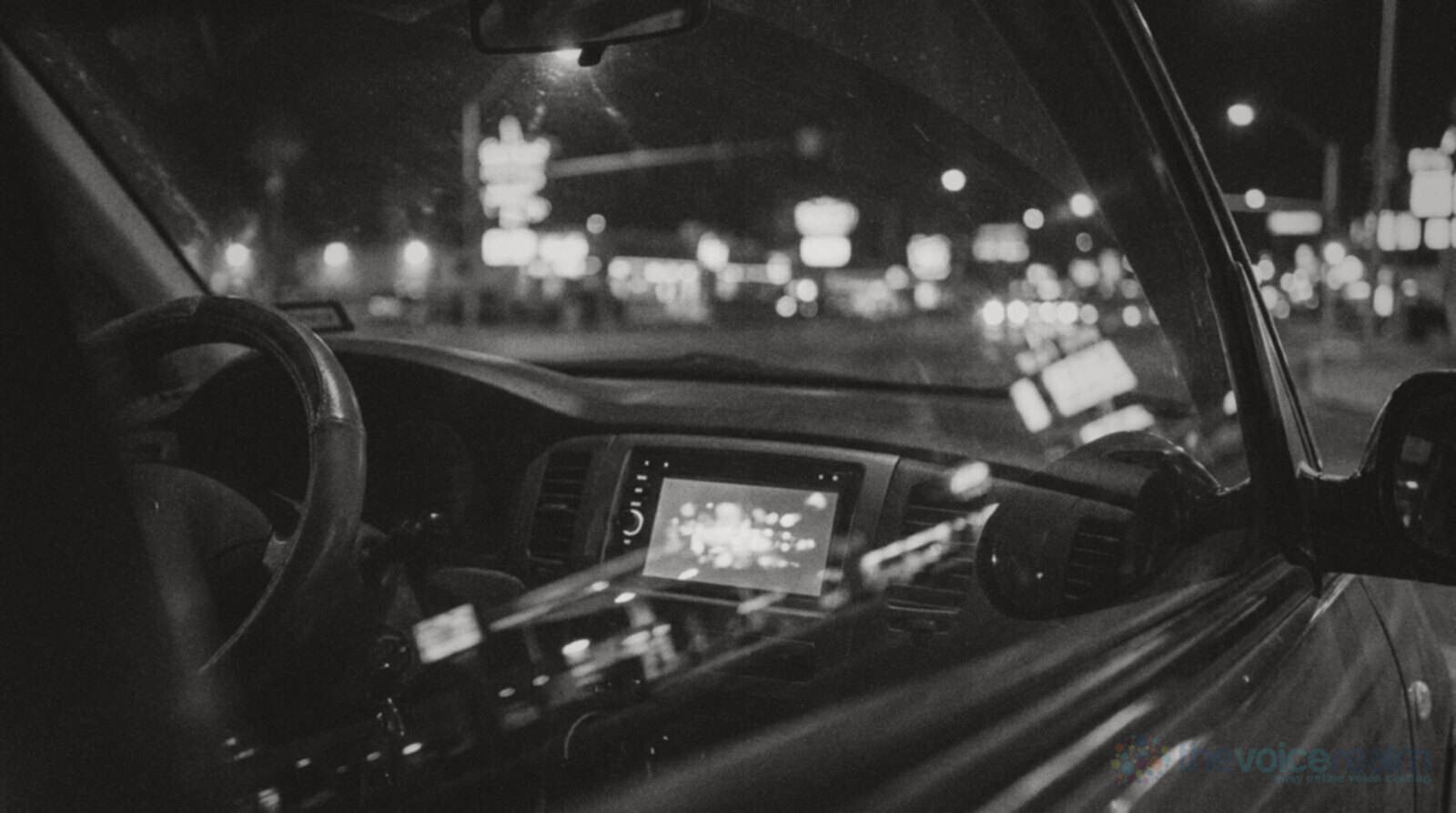 Inside a car at night, the dashboard display glowing, with the blurred neon lights of a city strip beyond the windscreen © The Voice Realm