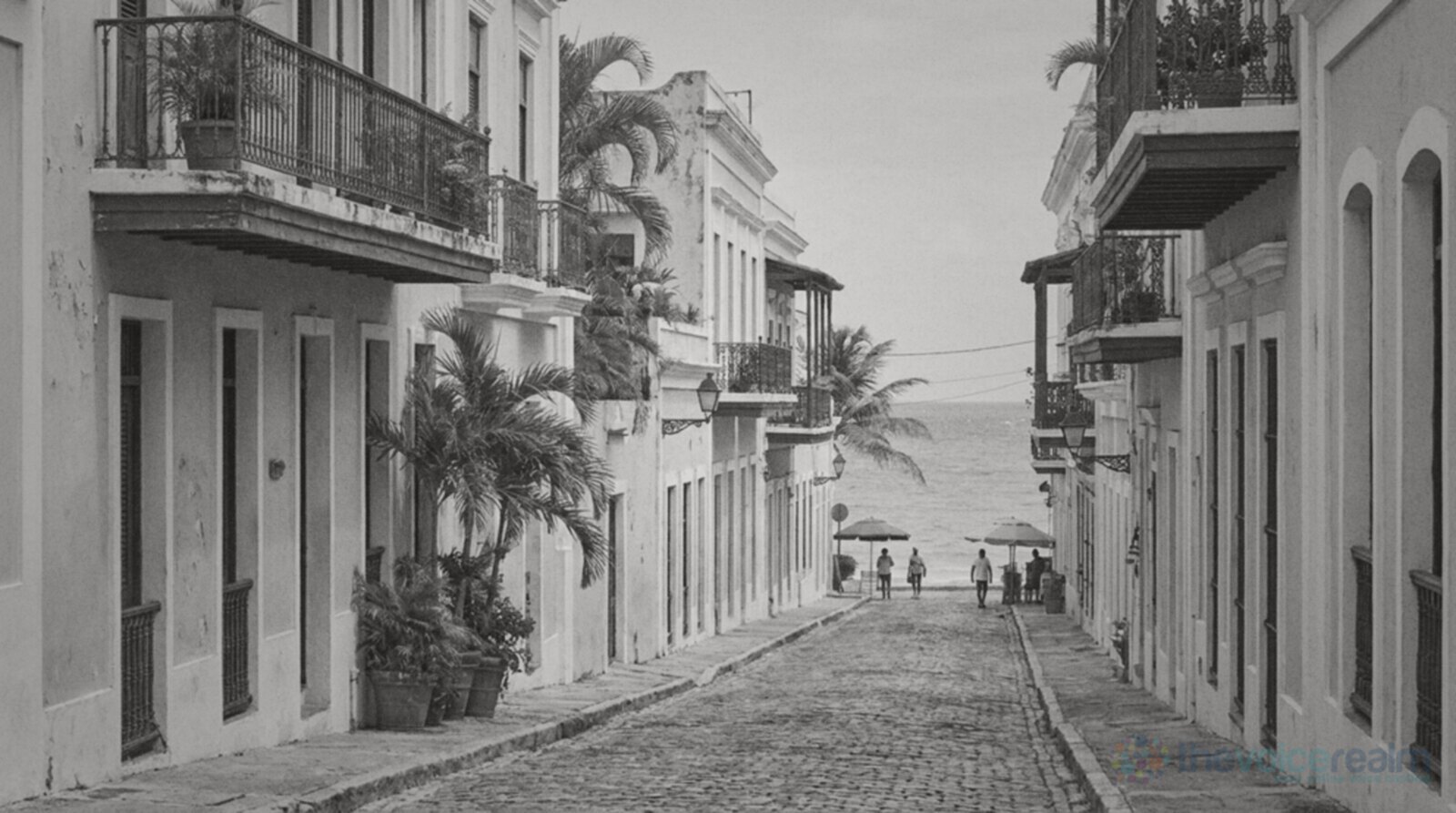 A cobblestone street of pastel colonial houses with balconies and palm trees leading down to a turquoise sea © The Voice Realm