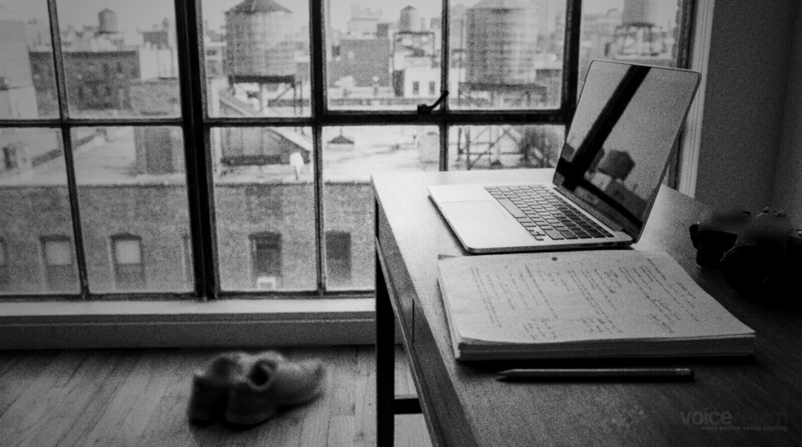 A desk by a big apartment window over city rooftops and water towers, with a laptop, a marked-up script, a camera and running shoes © The Voice Realm