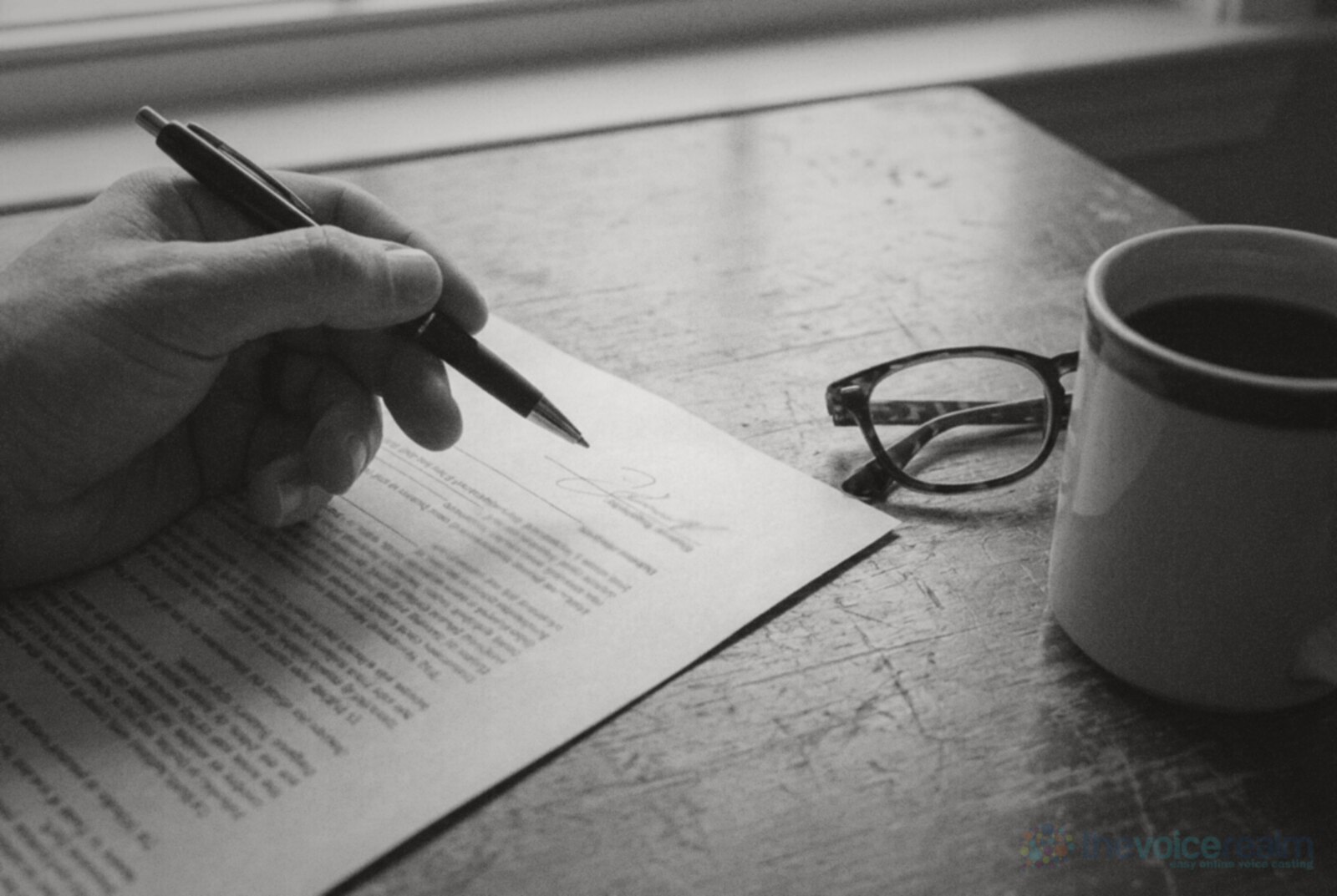A hand holding a pen over a printed contract on a wooden desk, with reading glasses and a mug of coffee © The Voice Realm