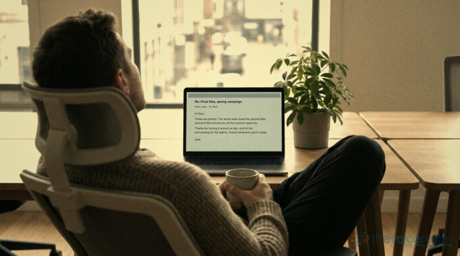 A relaxed client leaning back in an office chair with a coffee, looking at a laptop by a bright window © The Voice Realm