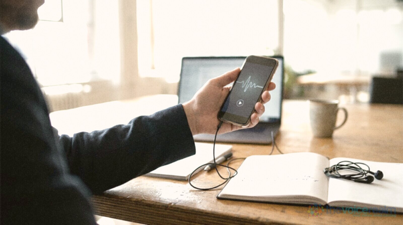 A person listening to short audio clips on a phone with wired earbuds, a ticked notebook on the desk © The Voice Realm