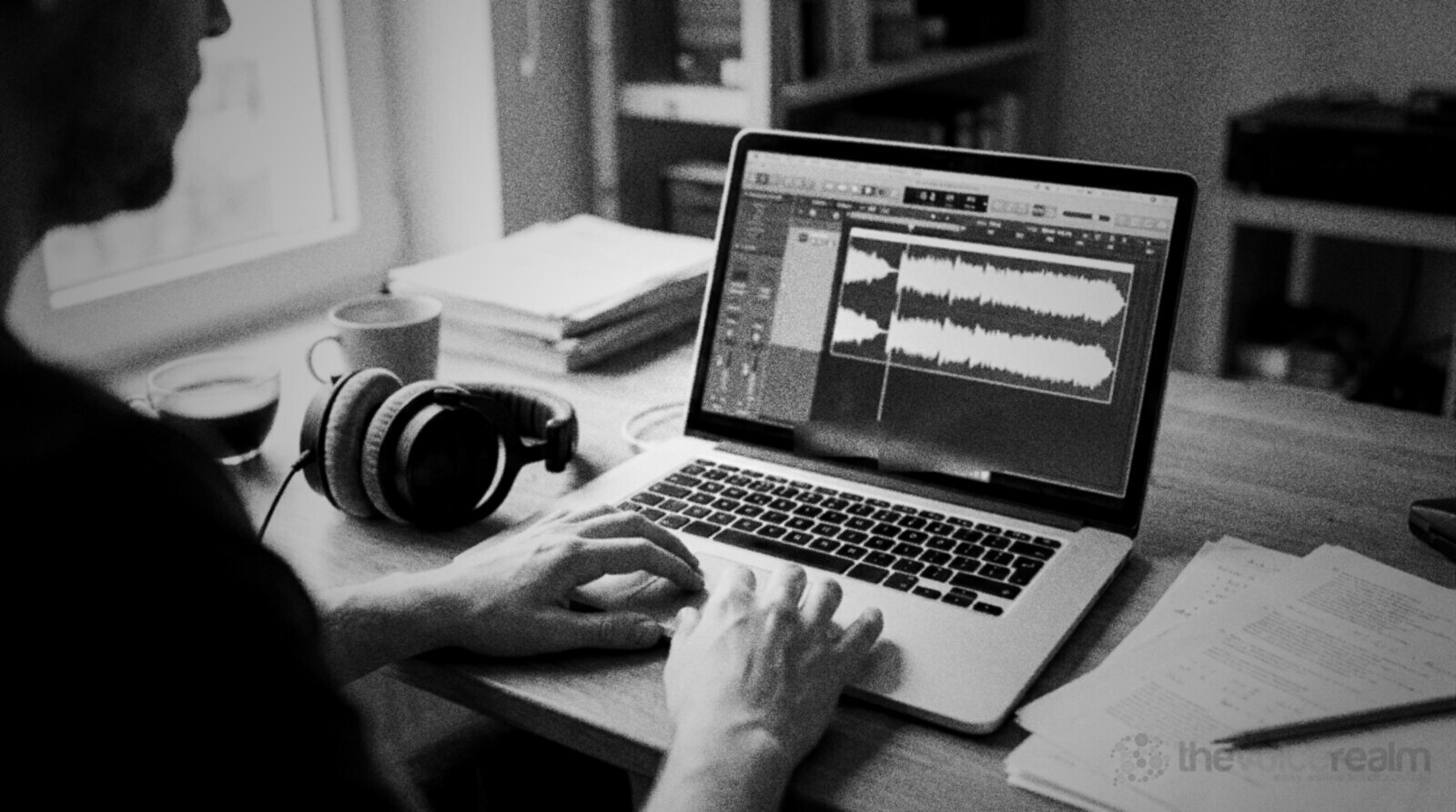 Hands on a laptop checking a waveform in an audio editor, headphones and a script on the desk beside it © The Voice Realm