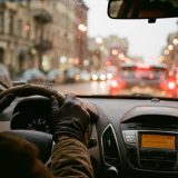 A driver's gloved hands on the steering wheel in slow evening city traffic, the dashboard radio glowing and red tail lights blurred through a rain-flecked windscreen © The Voice Realm