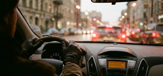 A driver's gloved hands on the steering wheel in slow evening city traffic, the dashboard radio glowing and red tail lights blurred through a rain-flecked windscreen © The Voice Realm