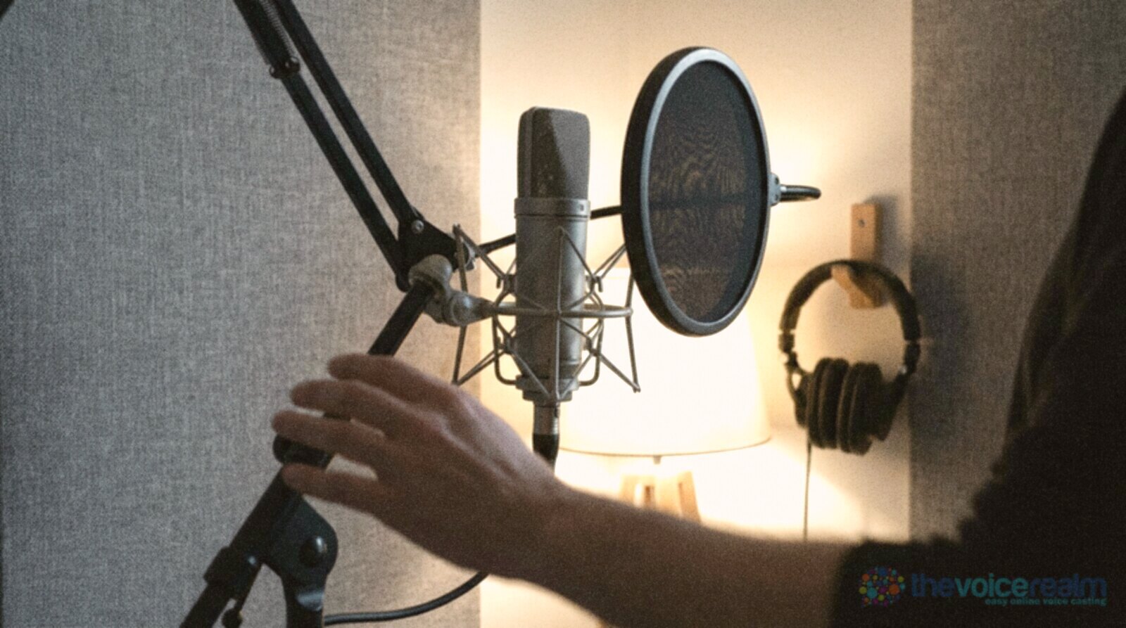 A studio condenser microphone in a shock mount on a boom arm with a round pop filter beside it, grey fabric panels, a warm lamp and headphones hanging on a hook © The Voice Realm