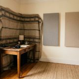 A spare bedroom corner set up for recording, a heavy woven blanket hung around a wooden desk with headphones on it, fabric acoustic panels on the wall and a full bookcase beside a jute rug © The Voice Realm