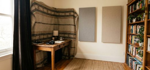 A spare bedroom corner set up for recording, a heavy woven blanket hung around a wooden desk with headphones on it, fabric acoustic panels on the wall and a full bookcase beside a jute rug © The Voice Realm