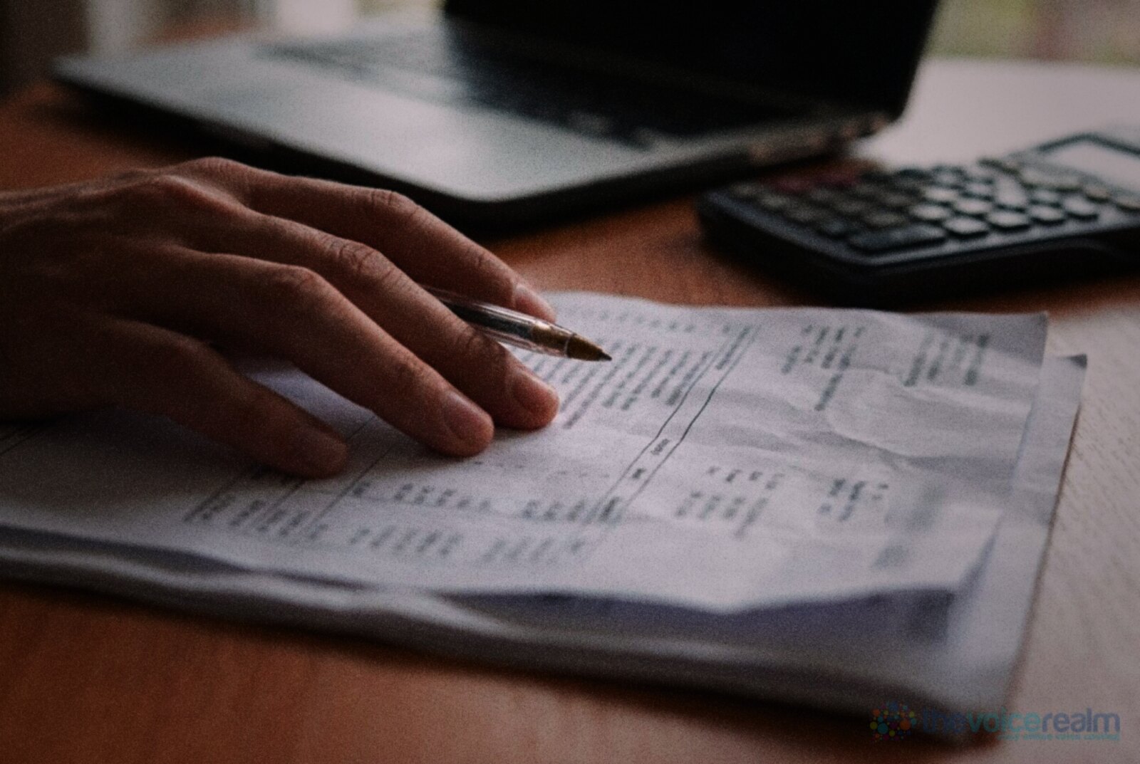 A hand resting a pen on a printed invoice beside a laptop and a calculator on a wooden desk © The Voice Realm
