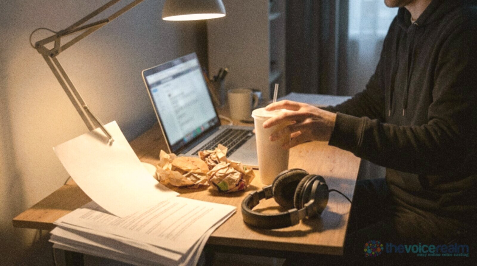 A home desk at night with burger wrappers, a soda cup, a laptop and headphones under a desk lamp © The Voice Realm