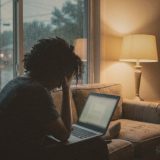 A person on a sofa at dusk with head in hand, looking at a laptop, rain on the window beside a lamp © The Voice Realm
