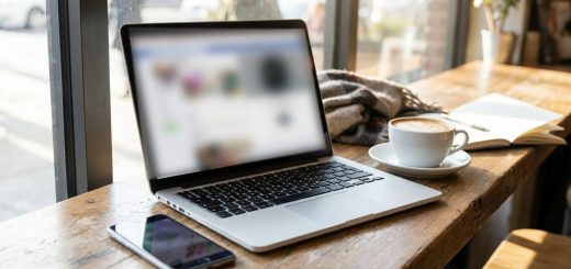 A laptop with a blurred feed on screen and a smartphone on a cafe table beside a flat white and an open notebook © The Voice Realm