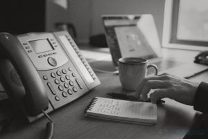 An office desk phone with its voicemail light on, beside a notepad of caller details and a coffee cup © The Voice Realm