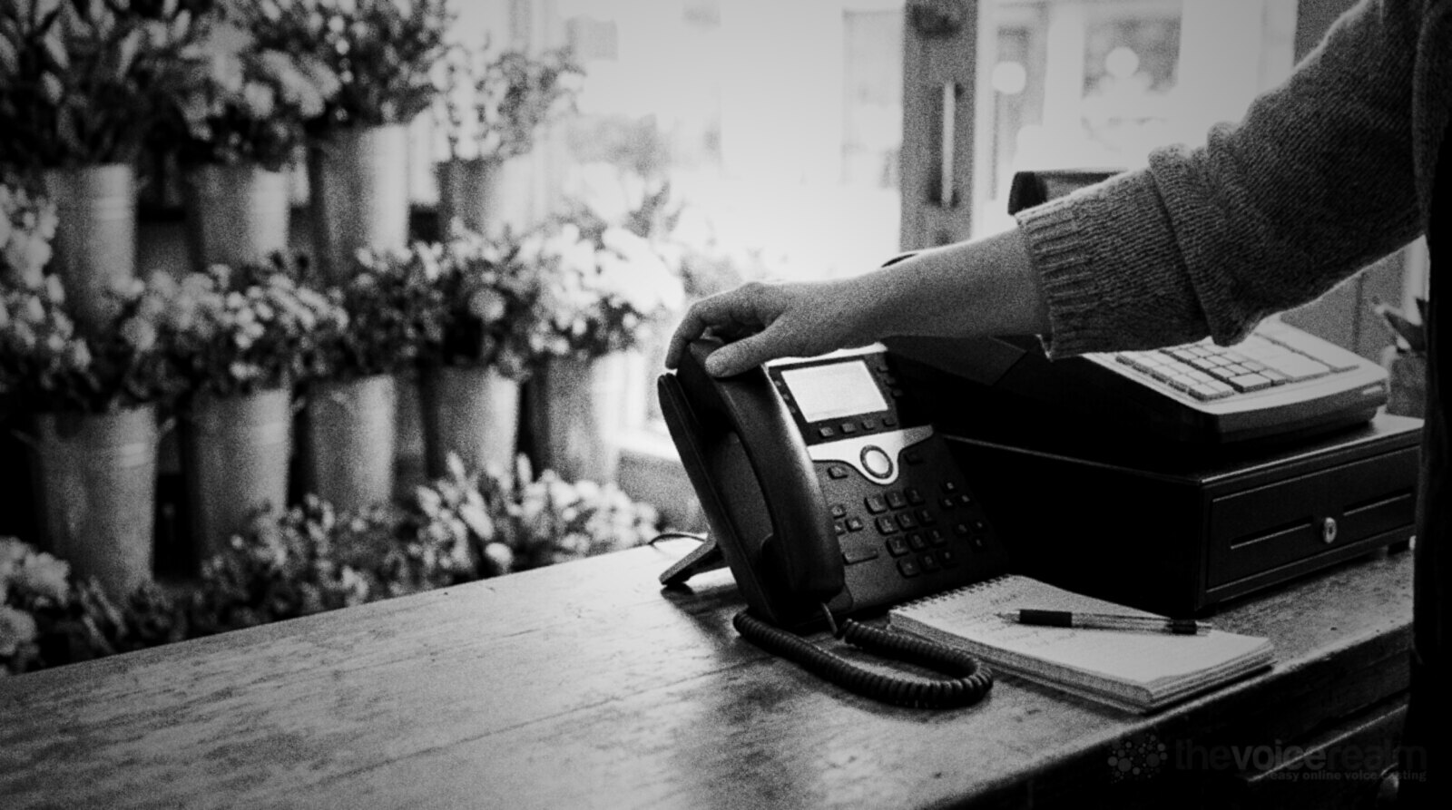 A shop owner's hand reaches for a desk phone on a florist's wooden counter, next to a till and a notepad © The Voice Realm
