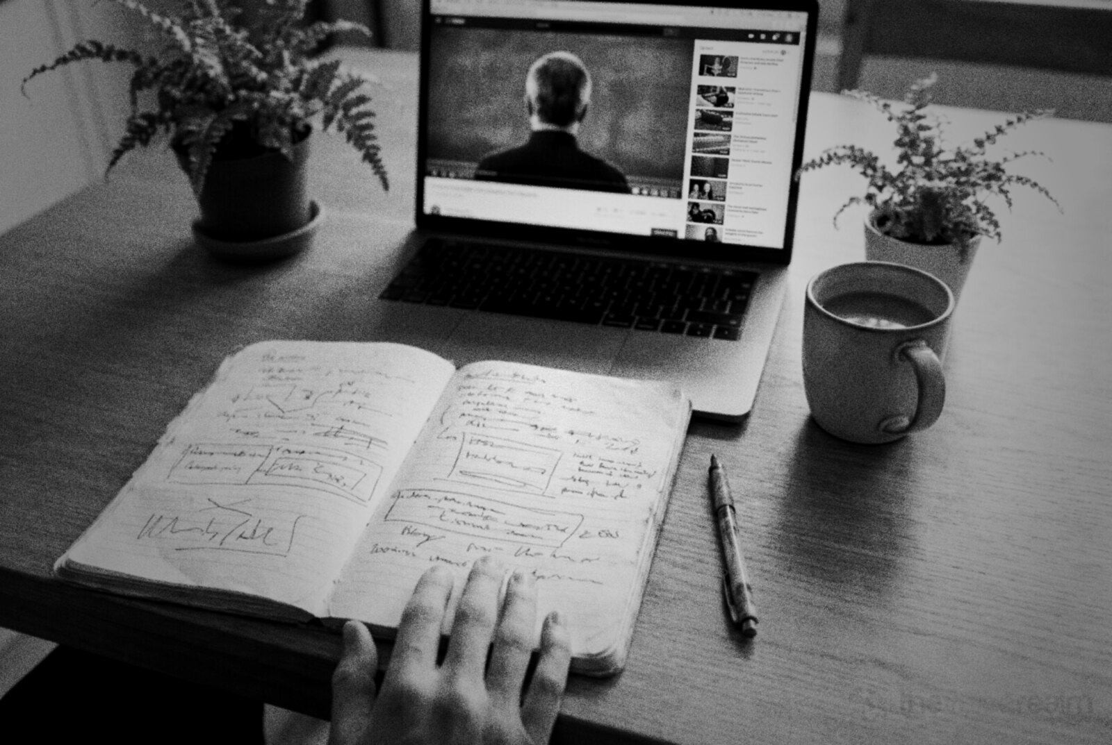 A hand on an open notebook of handwritten plans beside a laptop playing an online lecture and a mug of coffee © The Voice Realm