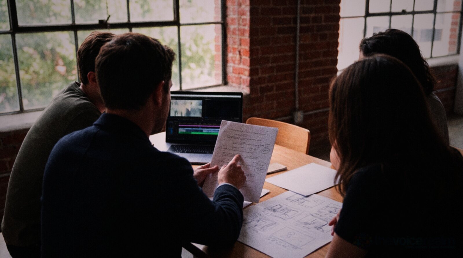 A small production team seen from behind around a table, one pointing at a marked-up script beside storyboard sketches and a laptop © The Voice Realm