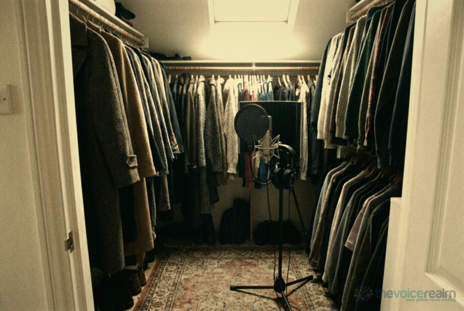 A walk-in wardrobe with coats and jumpers hanging densely on three sides and a studio microphone on a stand with a pop filter just inside the door © The Voice Realm
