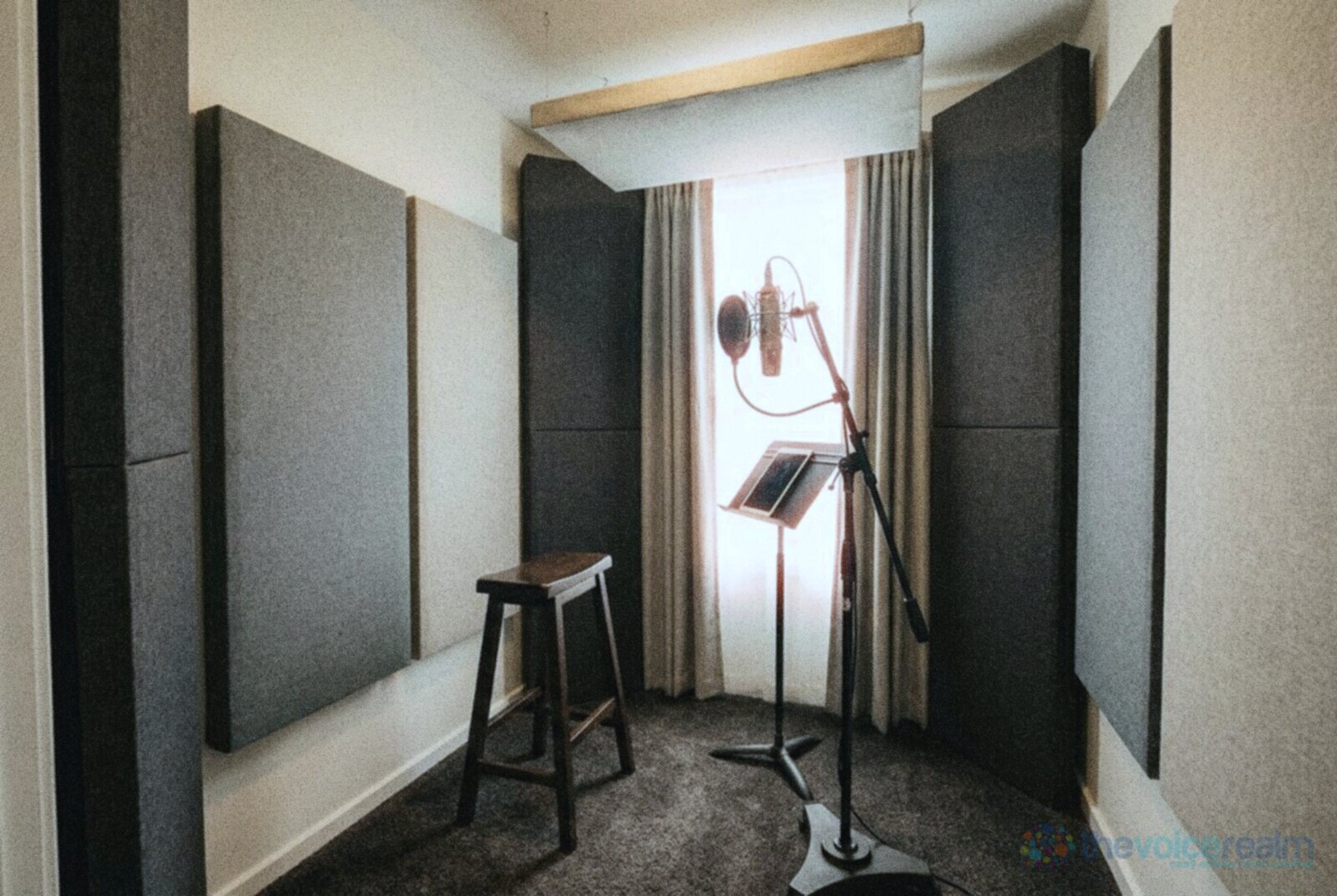 A small treated voice room with thick fabric panels on the walls, tall corner bass traps, a panel hanging from the ceiling and a microphone on a stand beside a stool © The Voice Realm