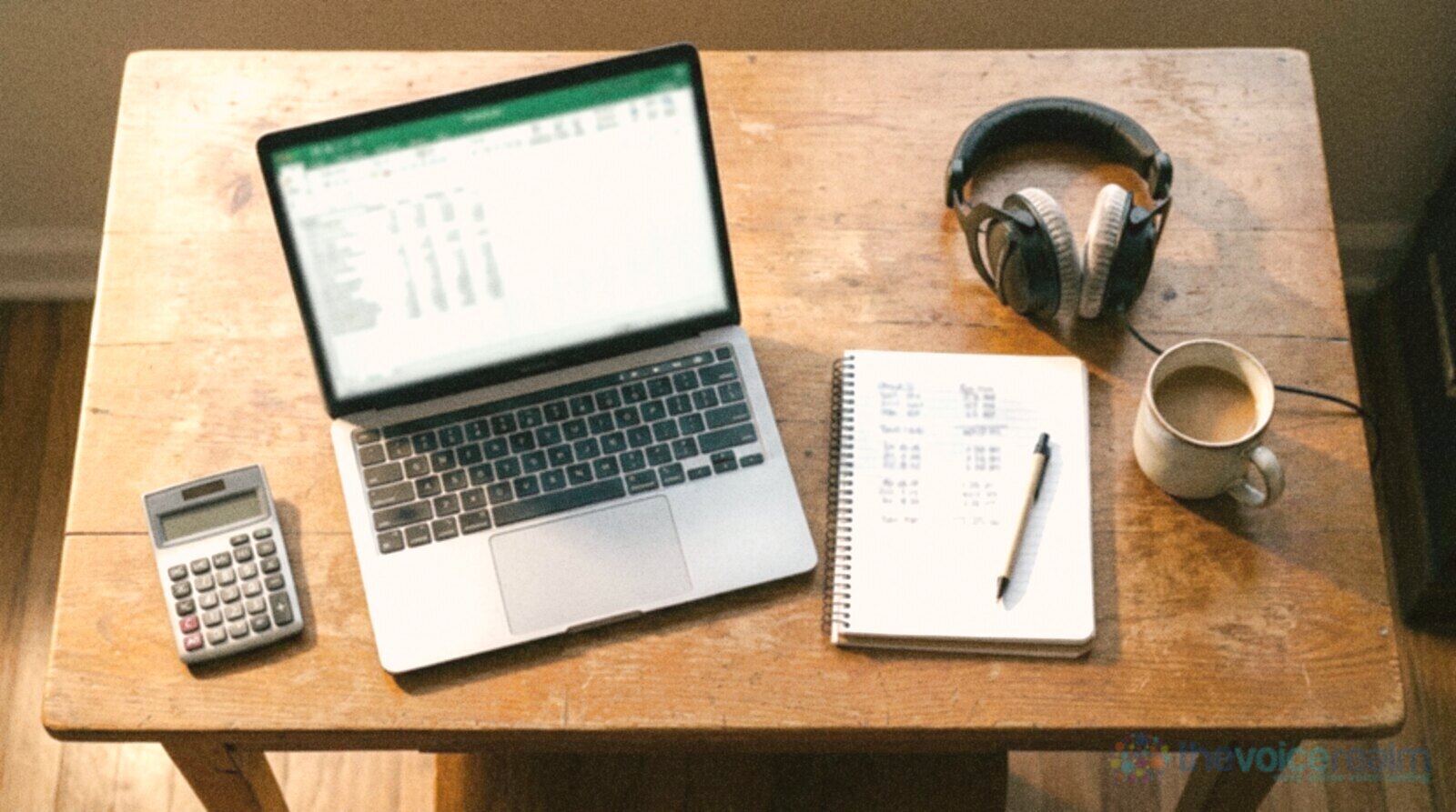 A wooden desk from above with a laptop spreadsheet, a calculator, a notebook of figures, studio headphones and a coffee © The Voice Realm