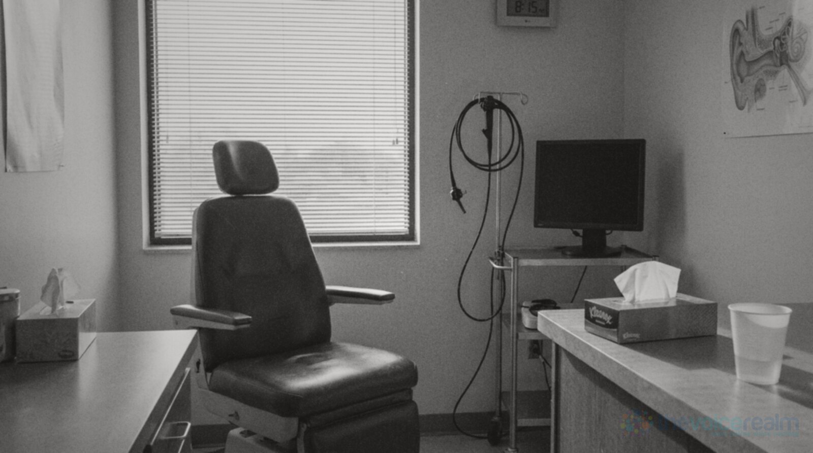 Black and white photo of an empty ear, nose and throat clinic room in the morning: a padded exam chair by a window with blinds, an endoscope coiled on a stand beside a switched off monitor, tissues and a paper cup of water on the counter © The Voice Realm
