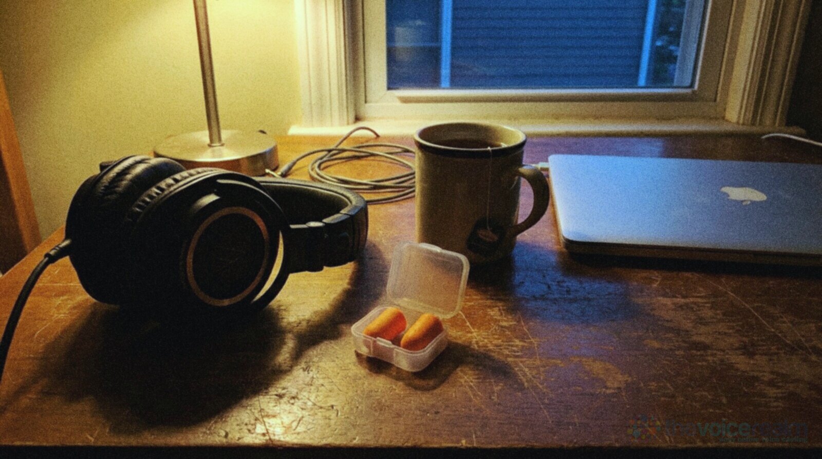 Closed-back headphones on a worn wooden desk at dusk beside a small open case of orange foam earplugs, a mug of tea and a closed laptop, lit by a desk lamp © The Voice Realm