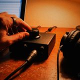 A hand turns the big gain knob on a small black two-input audio interface on a scuffed wooden desk at night, an XLR cable plugged into its front, closed-back headphones beside it and a laptop showing a recording session, in rich warm film colours © The Voice Realm