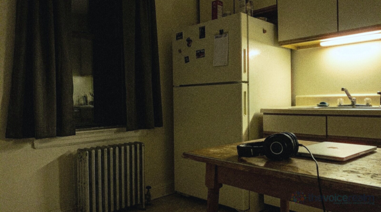 An empty home kitchen at night lit by a dim under-cabinet light, a tall white fridge in the corner, a radiator under a curtained window and closed-back headphones on a wooden table beside a closed laptop, in cool grainy night film colours © The Voice Realm